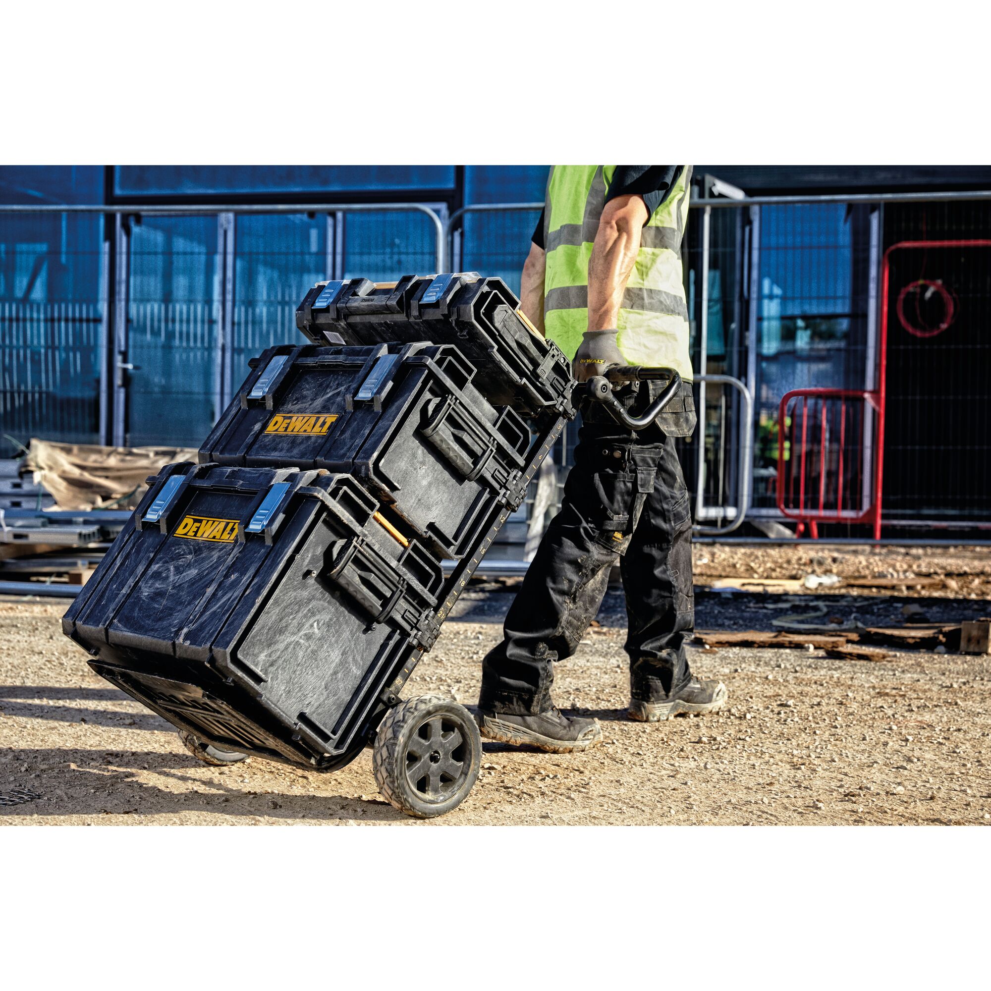 A person in work clothes and a safety vest is pulling a stack of three black DEWALT tool storage boxes on a wheeled trolley at a construction site.