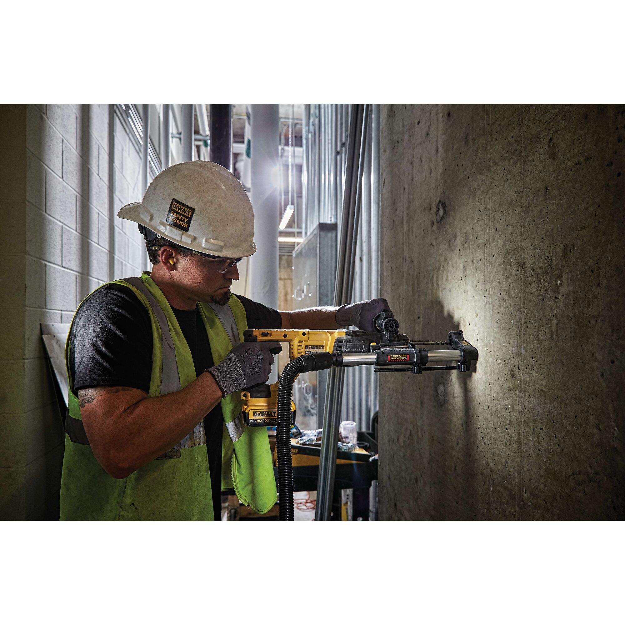 A construction worker wearing a hard hat and safety vest uses a DEWALT power tool with a dust extraction attachment to drill into a concrete wall at an indoor jobsite. The worker's face is blurred.