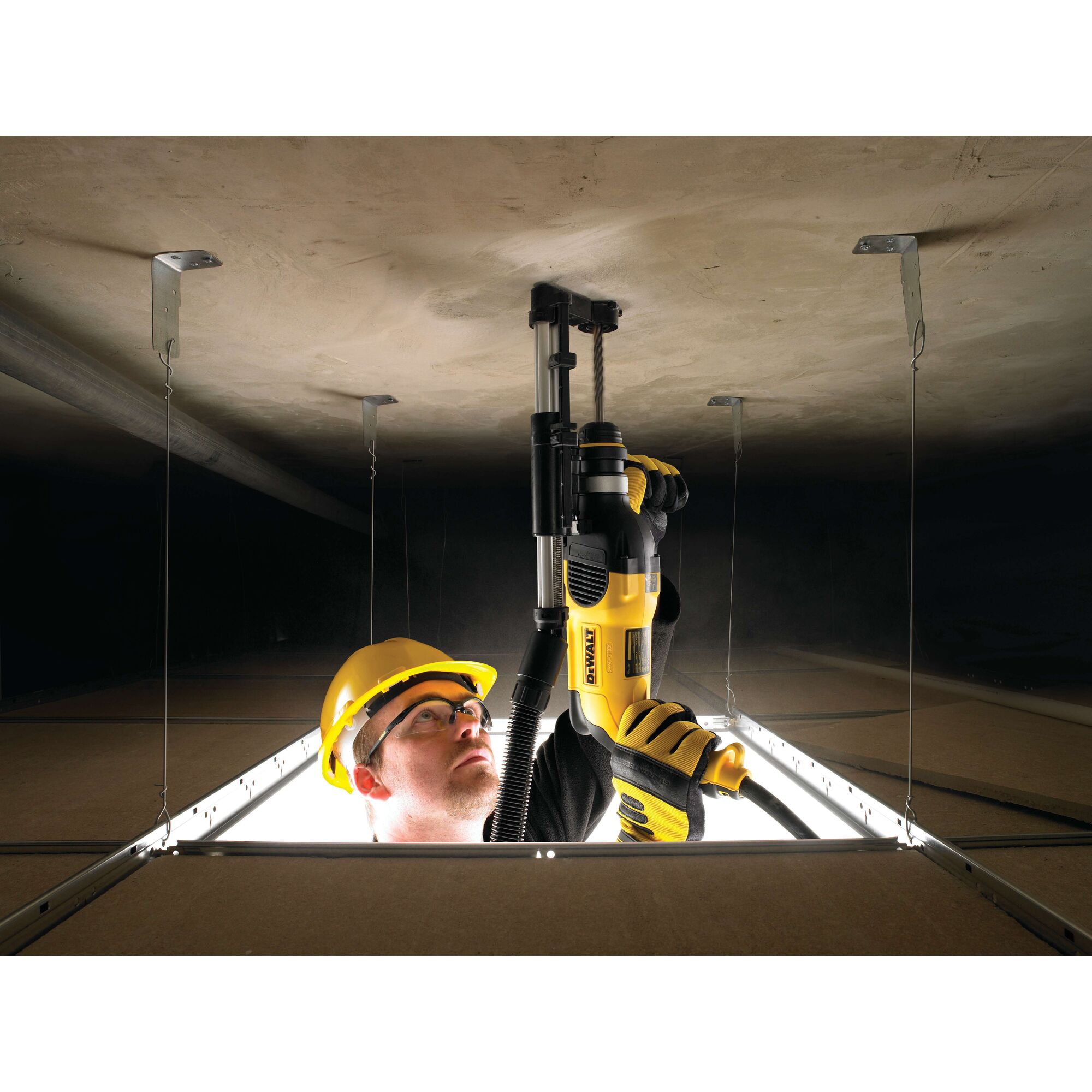 A person wearing a yellow safety helmet and gloves is using a DEWALT D25301D power tool to drill into a ceiling, seen from below through an open ceiling panel. The face of the person is blurred for privacy.