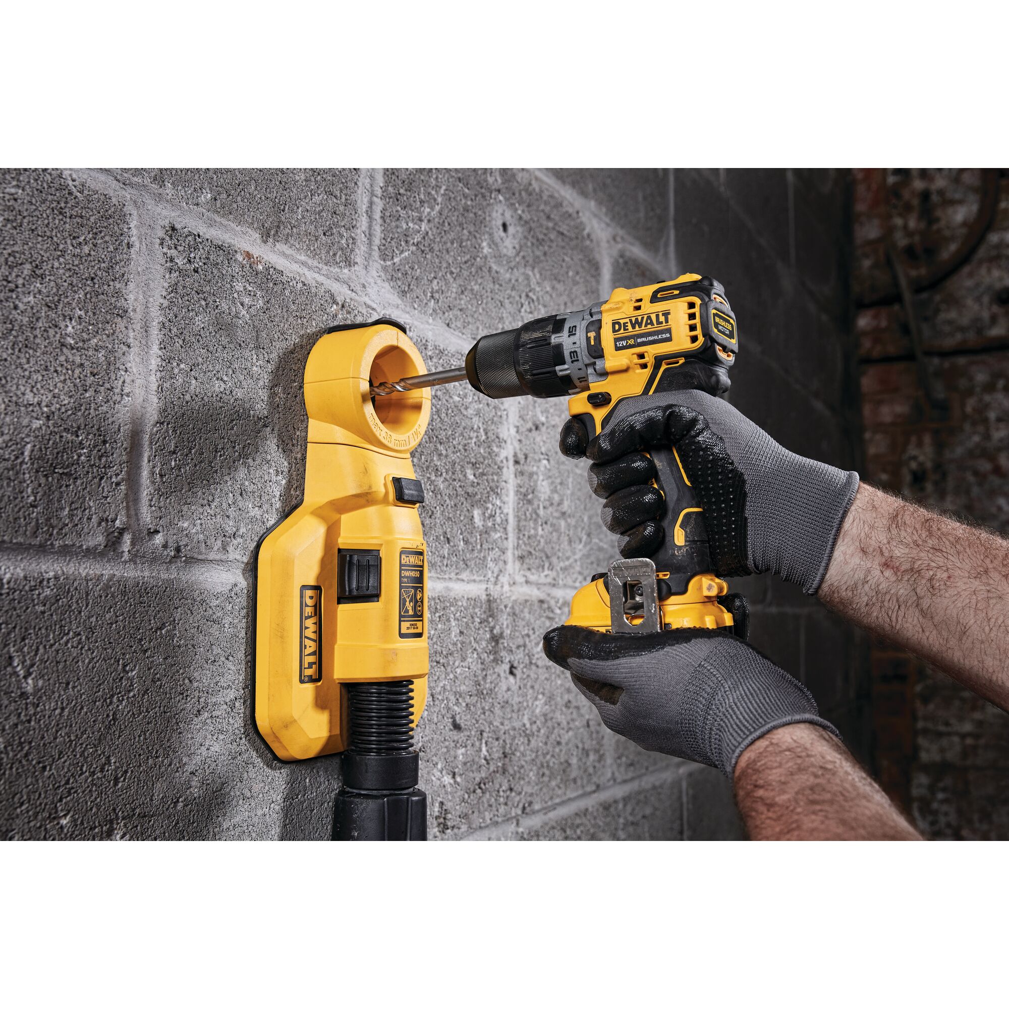 A person wearing gray gloves is using a yellow DEWALT cordless drill to drill into a concrete wall, with a yellow dust extractor attached to collect debris.