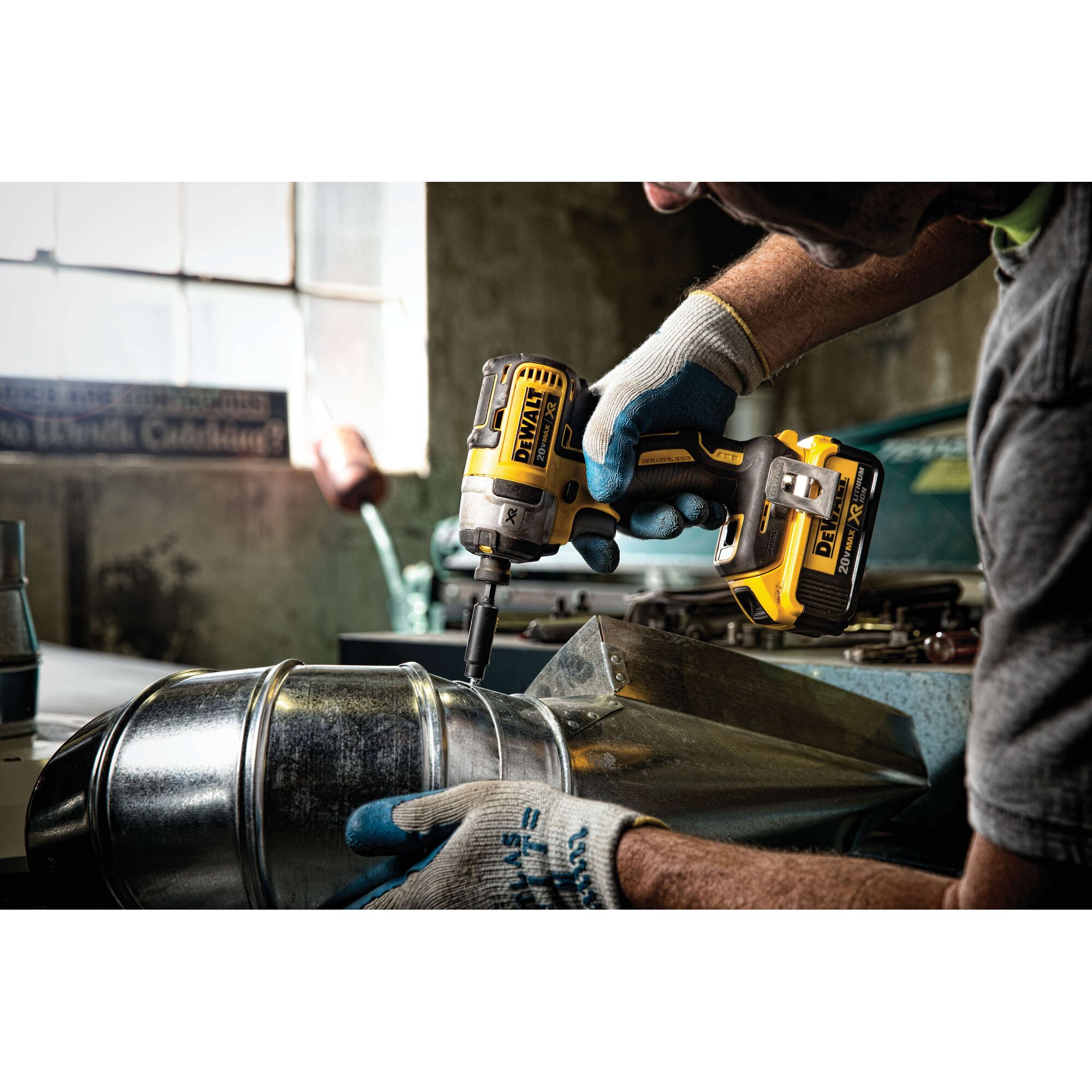 A person wearing work gloves is using a DEWALT DCF887M2 cordless impact driver to fasten a screw into a metal duct inside a workshop.