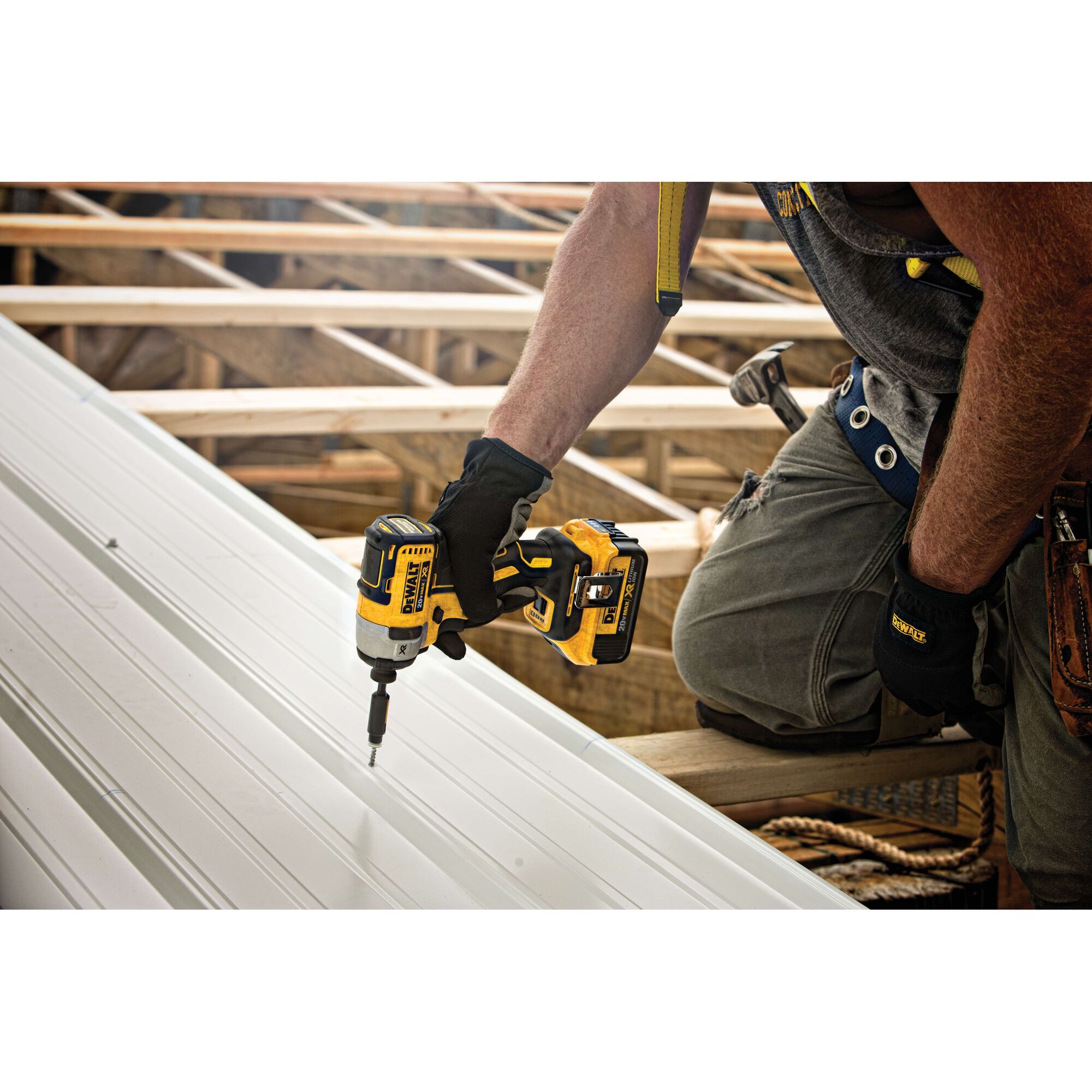A person wearing work gloves and a tool belt uses a yellow and black DEWALT cordless impact driver to fasten screws into a metal panel while working on a construction site with wooden framing.