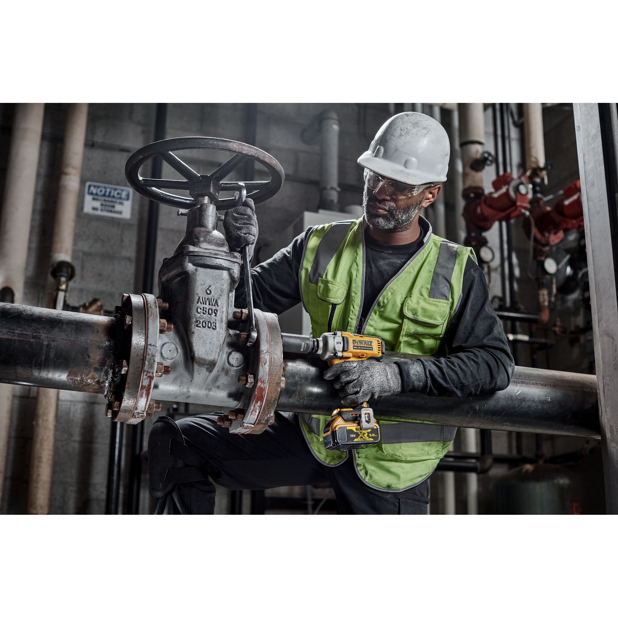 A worker wearing a green safety vest and helmet is using a DEWALT DCF891P2T power tool to work on a large industrial valve attached to metal pipes inside a facility.