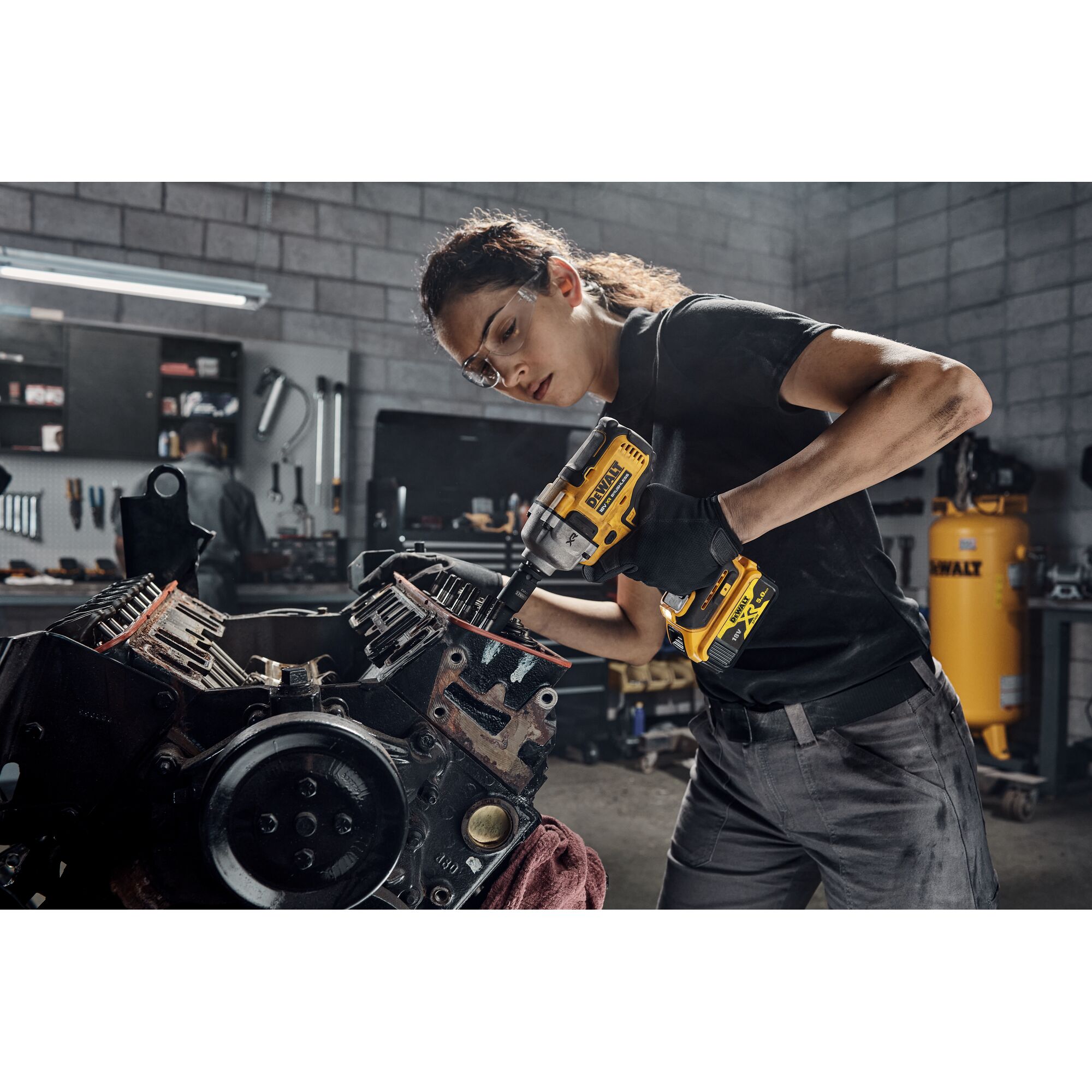 Person using a DEWALT DCF891P2T cordless impact wrench to work on an engine block in a workshop setting. The individual is wearing work gloves and the DEWALT equipment is prominently displayed.
