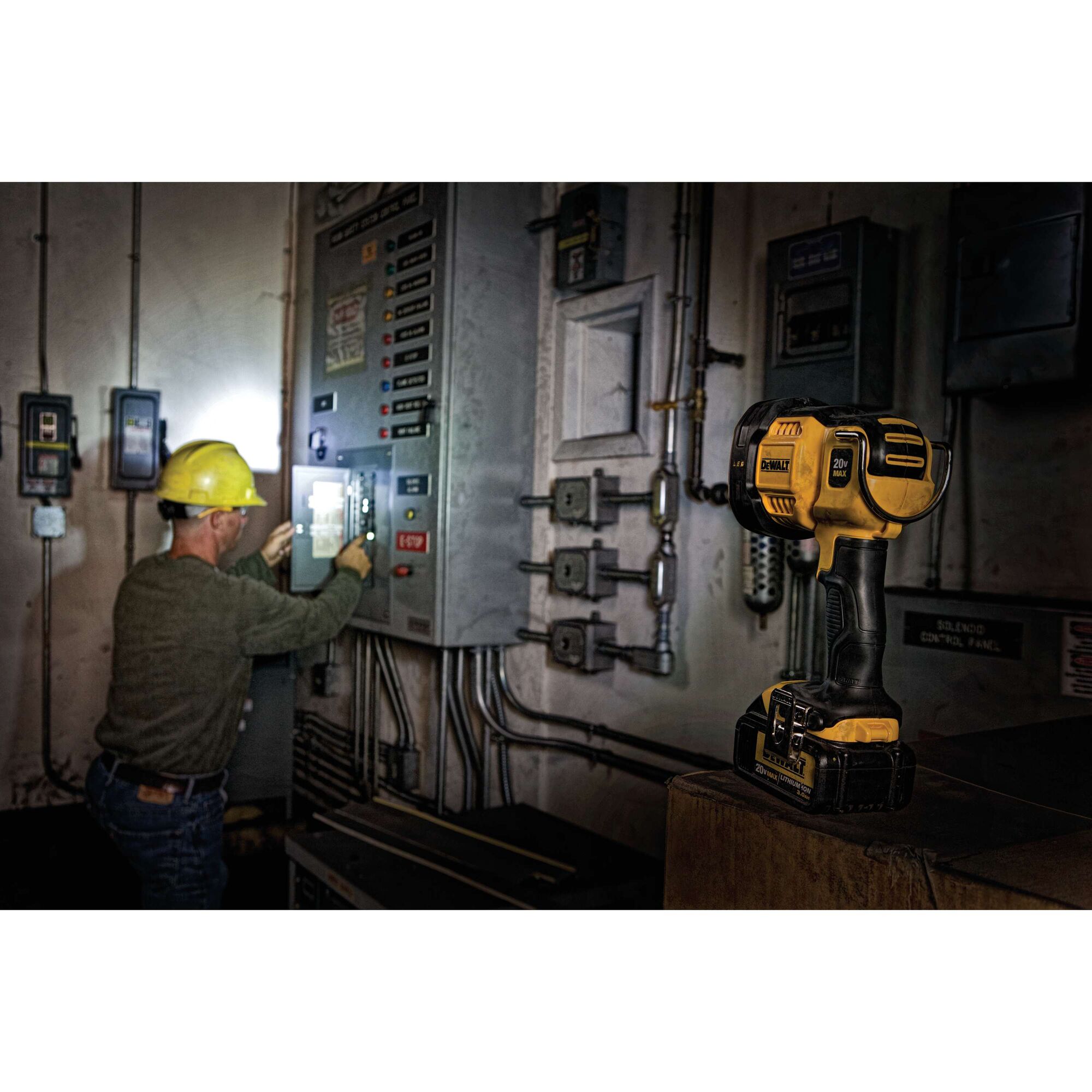 A DEWALT DCL043 LED work light is shown resting on a surface in a dimly lit industrial setting. In the background, a person wearing a yellow hard hat is working with electrical control panels, illuminated by the work light.