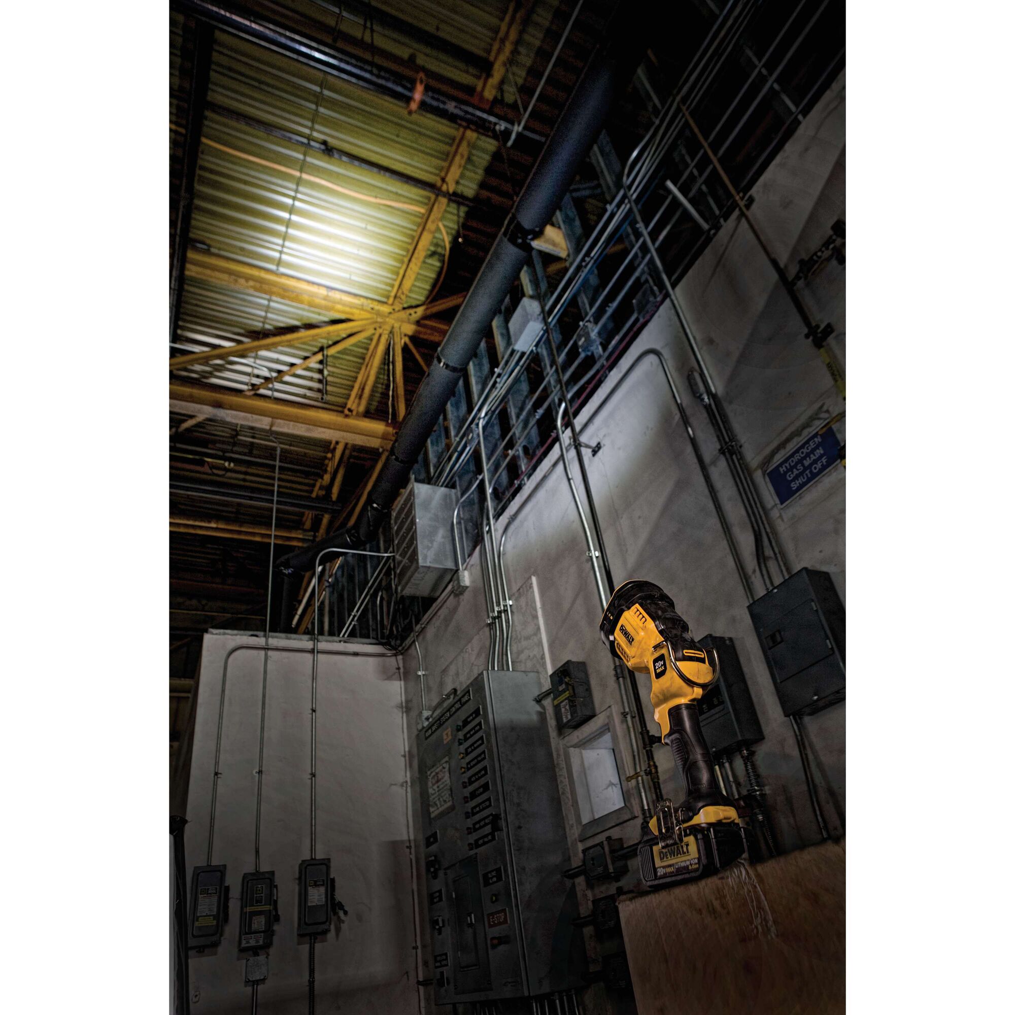 A DEWALT DCL043 handheld LED work light illuminating the ceiling of an industrial electrical room, with various electrical panels and conduit visible.