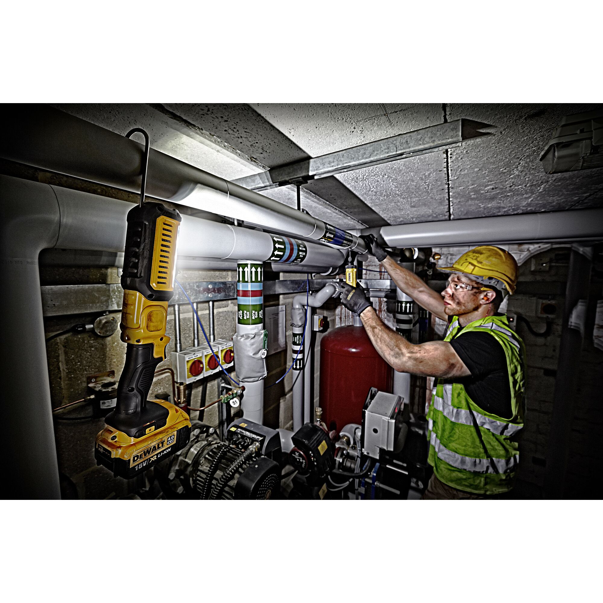 A worker wearing a safety vest and helmet is installing or repairing pipes in an industrial setting. A DEWALT DCL050 portable work light is illuminating the workspace.
