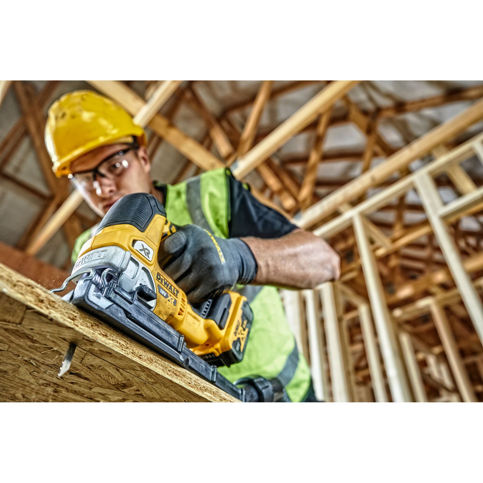 A person wearing construction safety gear uses a DEWALT DCS334N cordless jigsaw to cut a wooden board inside a building under construction.
