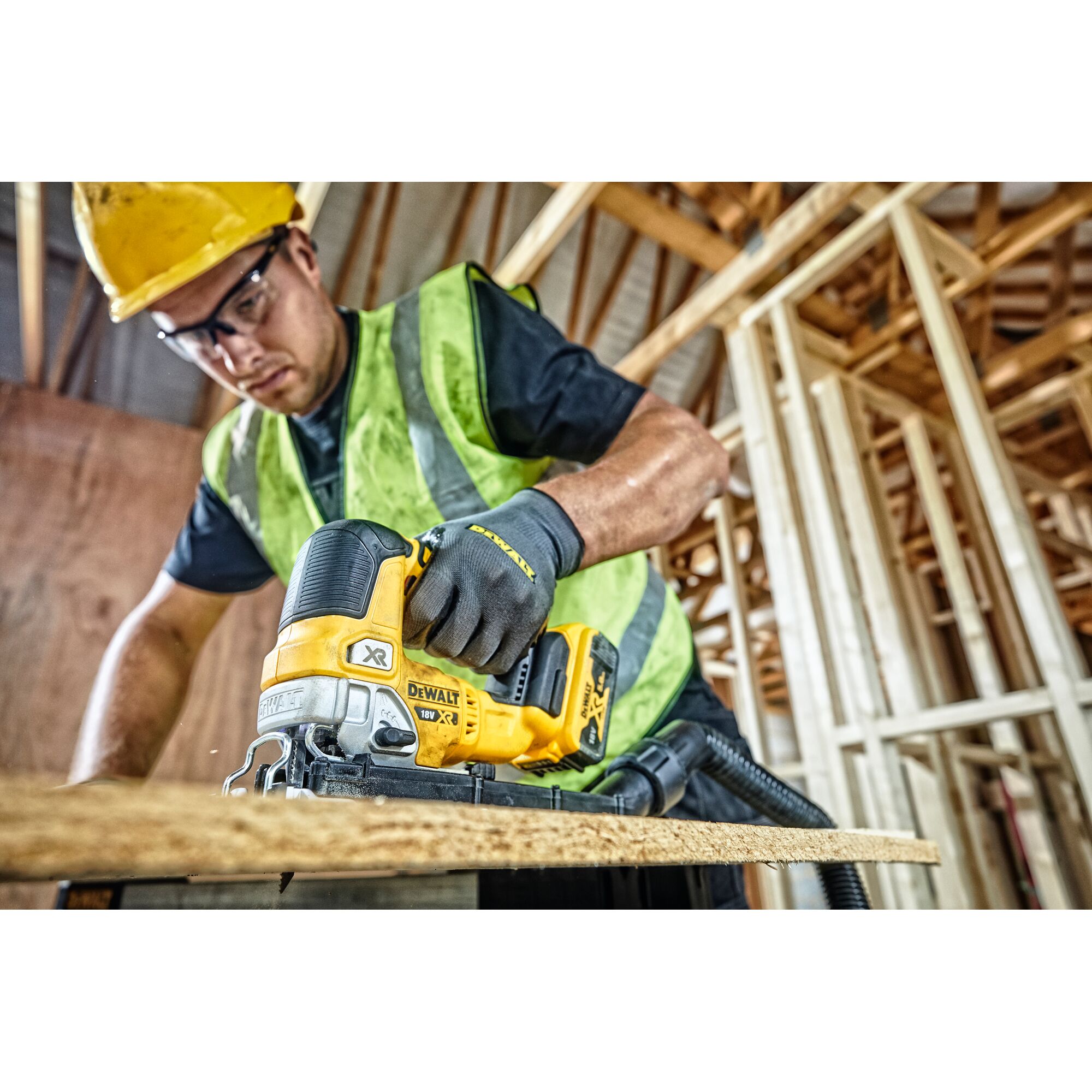 A construction worker wearing a yellow safety helmet and green vest is using a DEWALT DCS334N cordless jigsaw to cut a piece of wood, inside a building structure under construction.