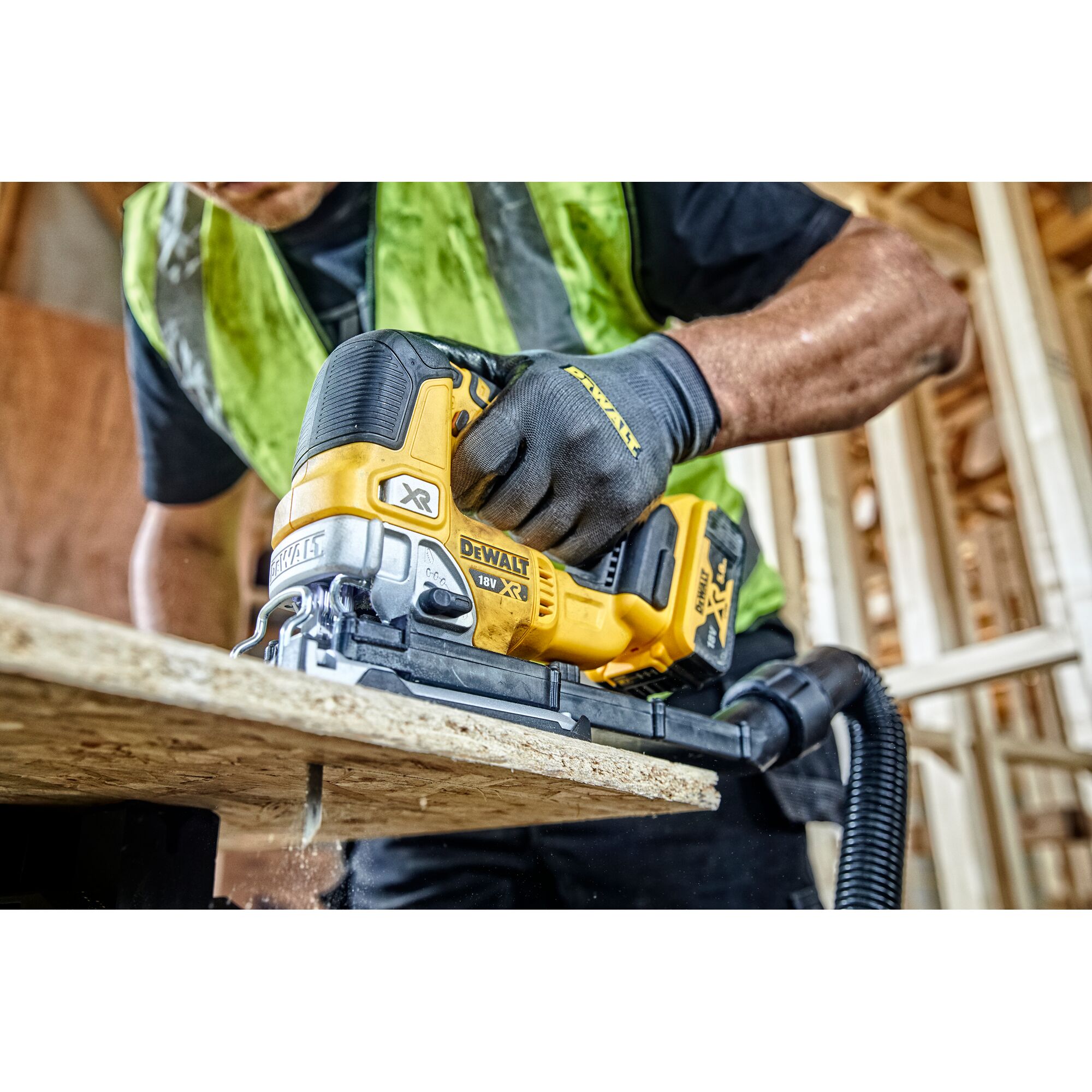 A person wearing a safety vest and gloves is using a yellow DEWALT cordless jigsaw (model DCS334N) to cut a plywood board in a construction setting.