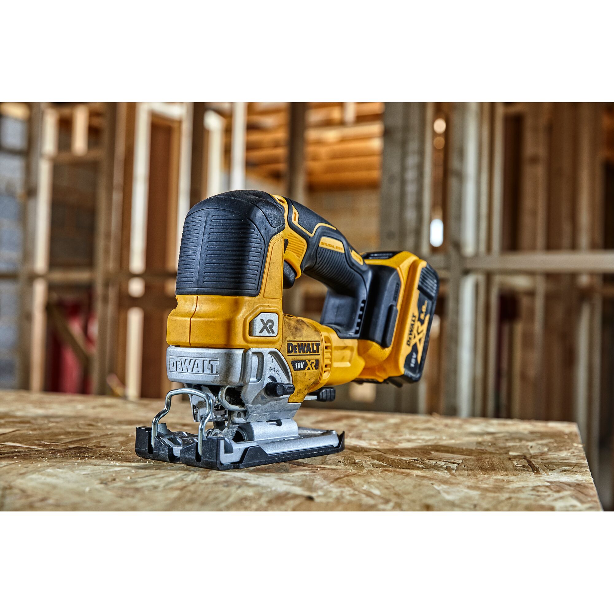 A DEWALT DCS334N cordless jigsaw, yellow and black, is placed on a wooden work surface inside a construction site with wooden framing in the background.