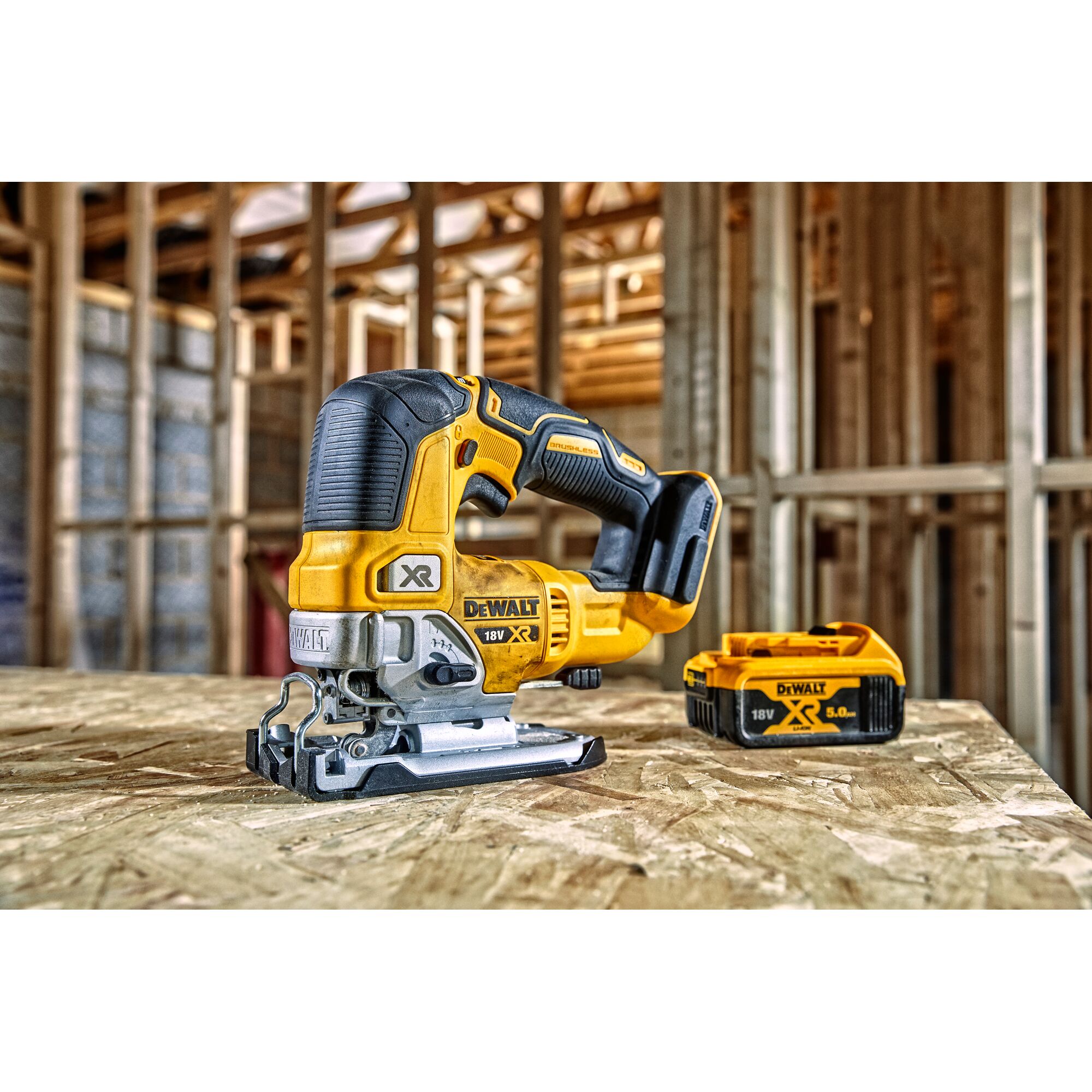 A DEWALT 18V XR cordless jigsaw with a yellow and black design is placed on a workbench inside a wooden construction site. A DEWALT 18V XR 5.0Ah battery is positioned next to the tool.