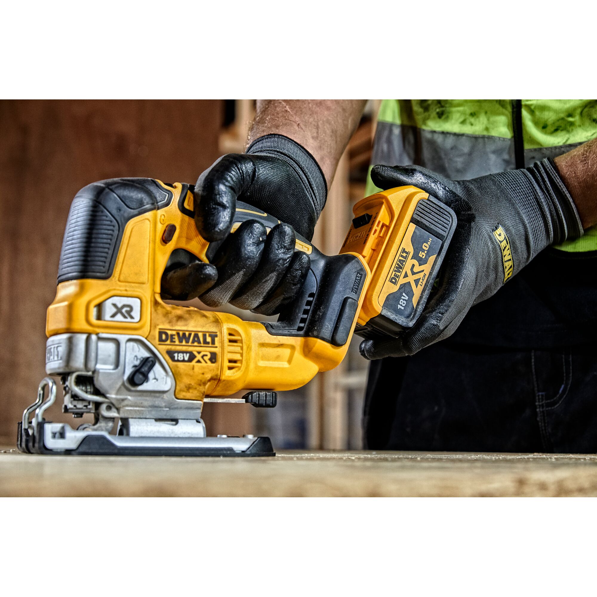 A close-up view of a person wearing gloves and holding a yellow DEWALT 18V XR cordless jigsaw, model DCS334N, in an indoor workshop environment.