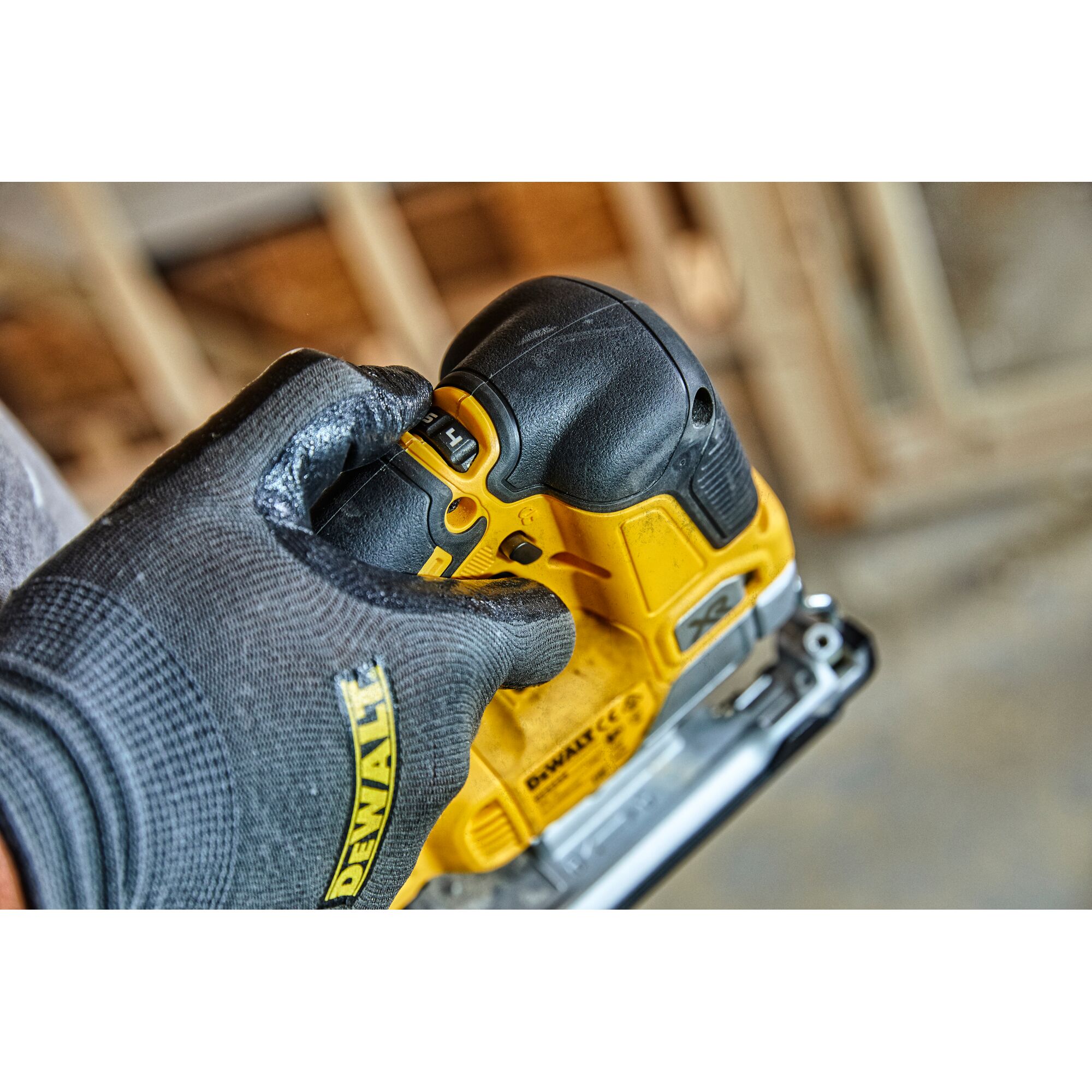 A close-up view of a person wearing a DEWALT-branded glove adjusting a setting on a yellow and black DEWALT power tool, possibly a cordless jigsaw, in a workshop environment.