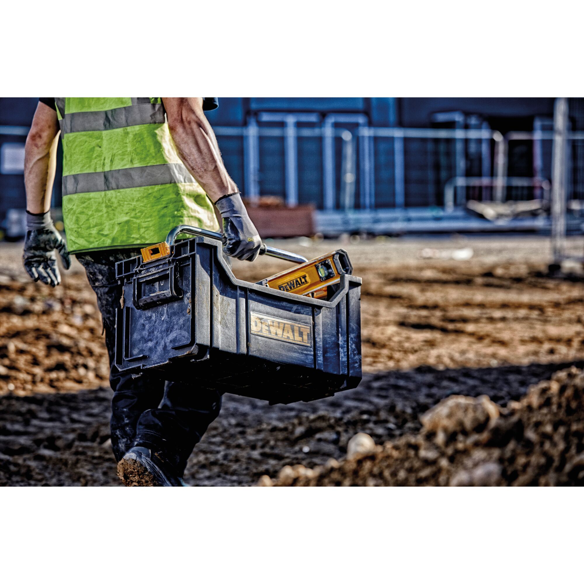 A person wearing a high-visibility vest and gloves carries a DEWALT tool box with tools, including a yellow spirit level, on a construction site with dirt ground and fencing in the background.