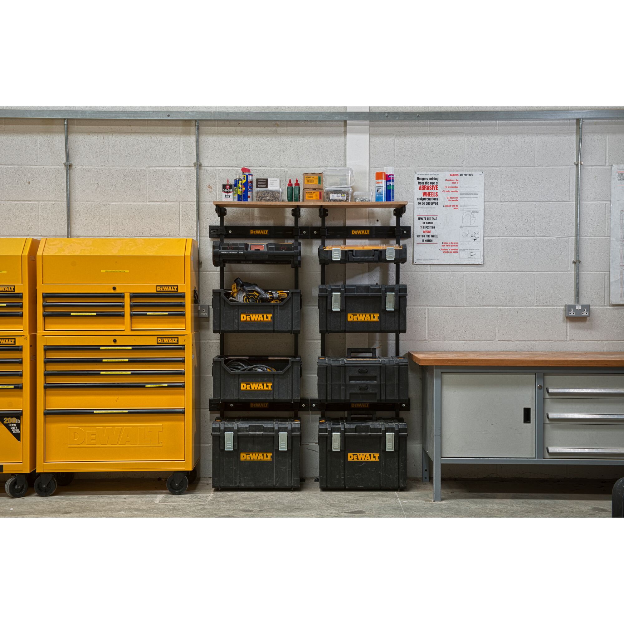 A workshop scene featuring DEWALT storage units, including yellow tool chests on the left and black modular toolboxes stacked on shelves in the center. The workspace is organized with tools, equipment, and supplies visible. A workbench with drawers is situated on the right, against a light gray wall.