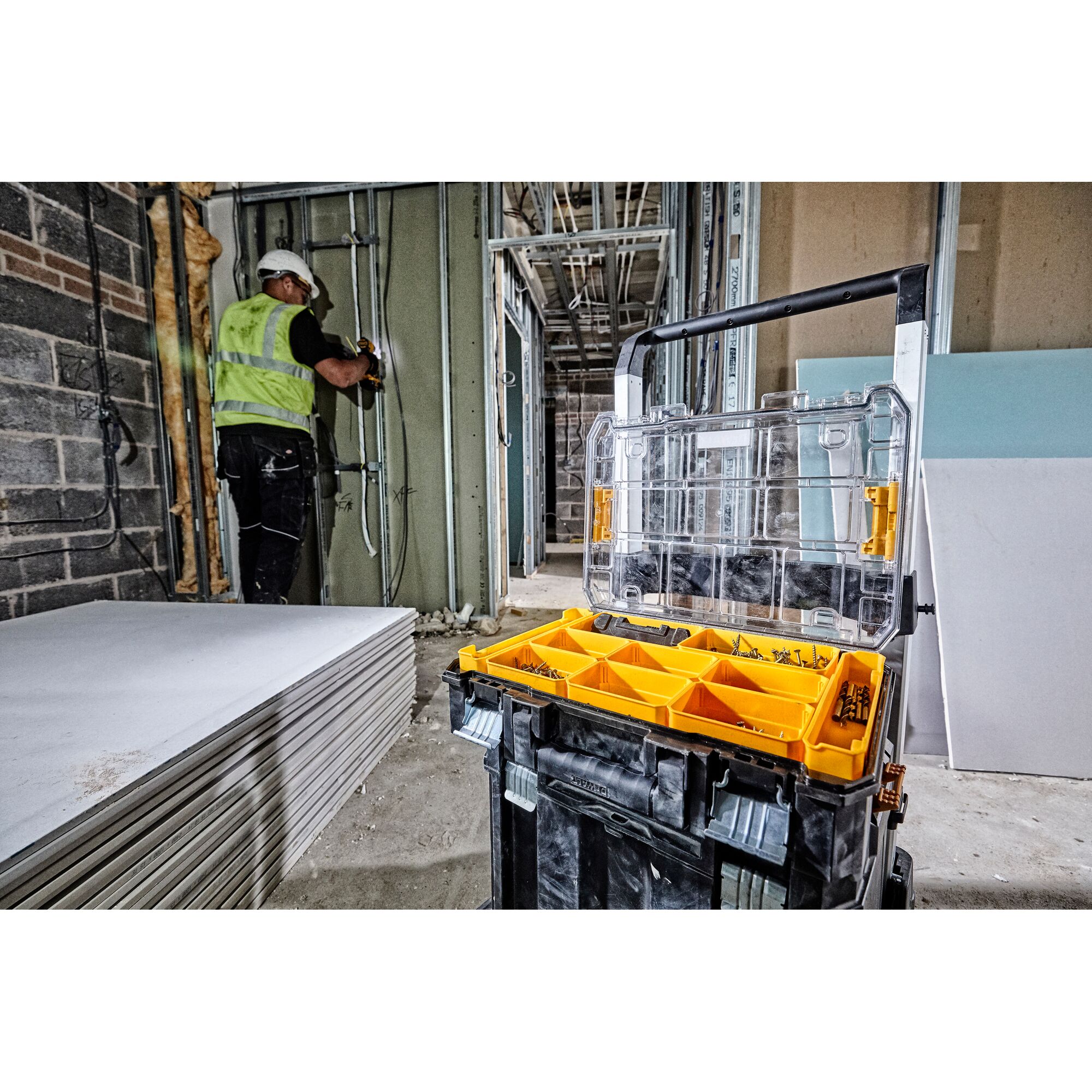 A DEWALT tool organizer box with yellow compartments containing screws and fasteners is open in the foreground on a construction site. In the background, a construction worker wearing a helmet and safety vest works on wiring inside a partially built wall. Sheets of drywall are stacked to the left.