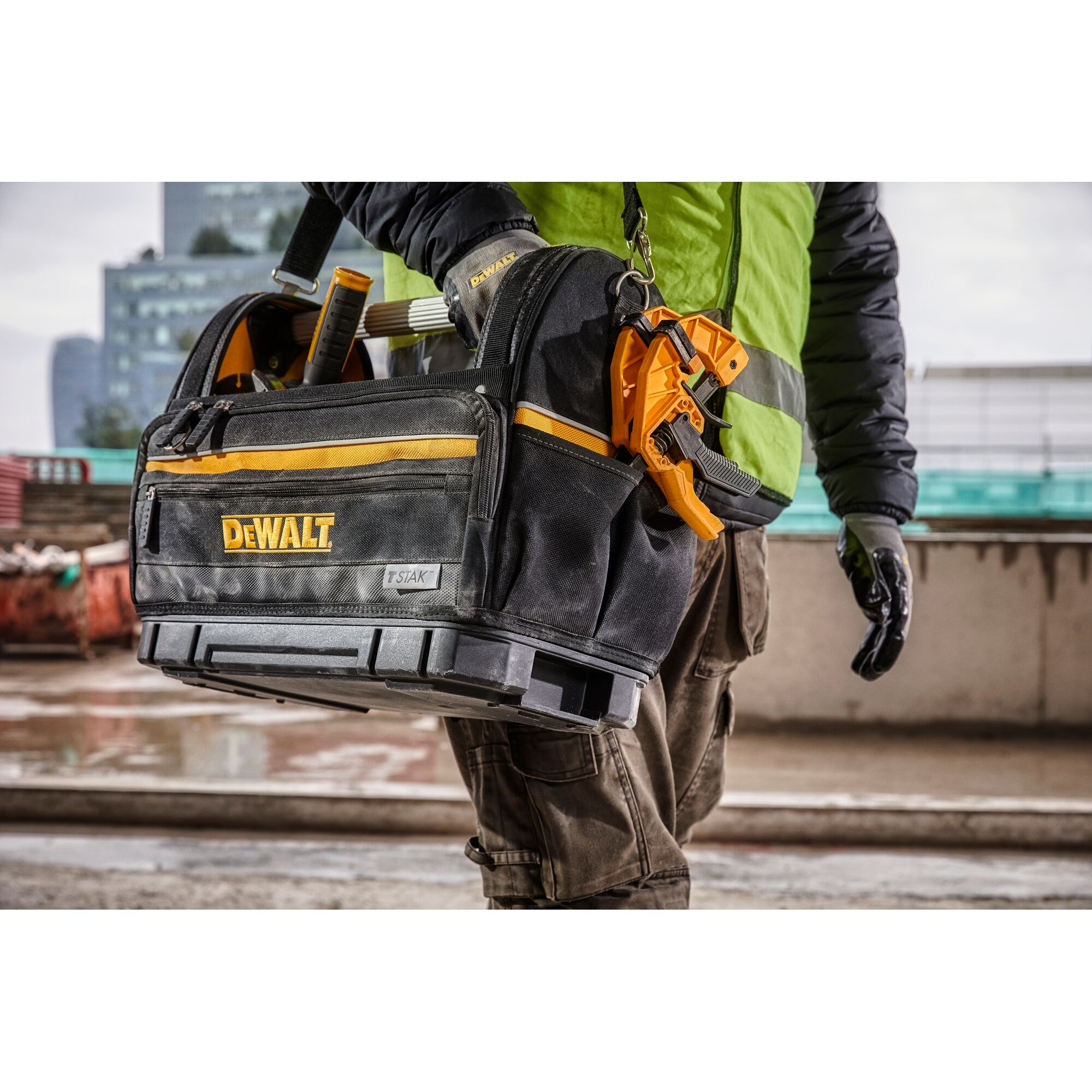 A DEWALT branded tool bag being carried by a person wearing work gloves and a high-visibility vest, with various tools visible inside the bag at a construction site.
