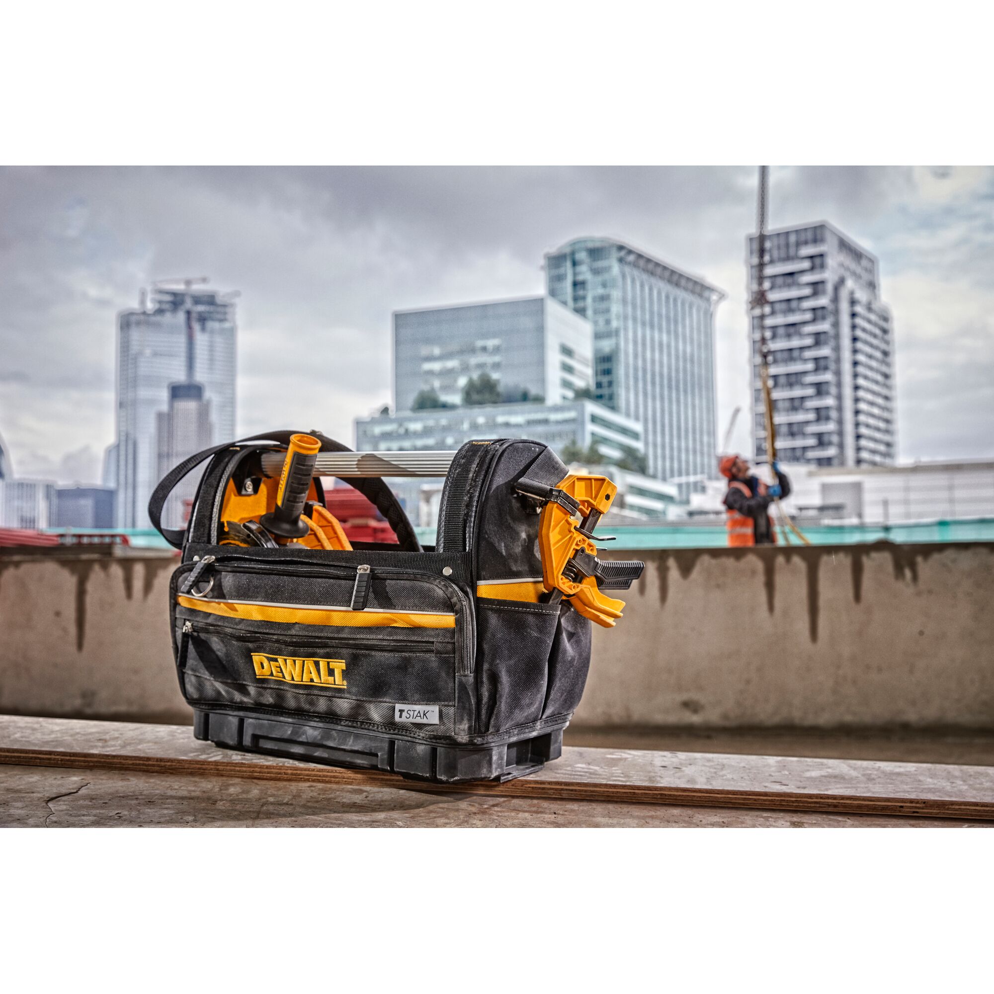 A DEWALT branded tool bag containing various tools, including clamps and a tape reel, is placed on a construction site with modern city buildings visible in the background.
